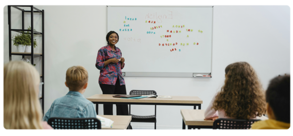 A young teacher stands at a marker board at the front of a class of students.