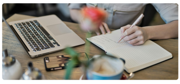 A student sits in front of a laptop next to a cup of coffee and writes in a journal.